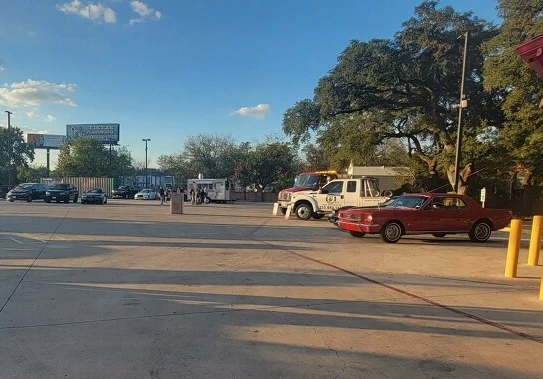 People sit in folding chairs in front of classic cars at a car show, under a blue sky with clouds.